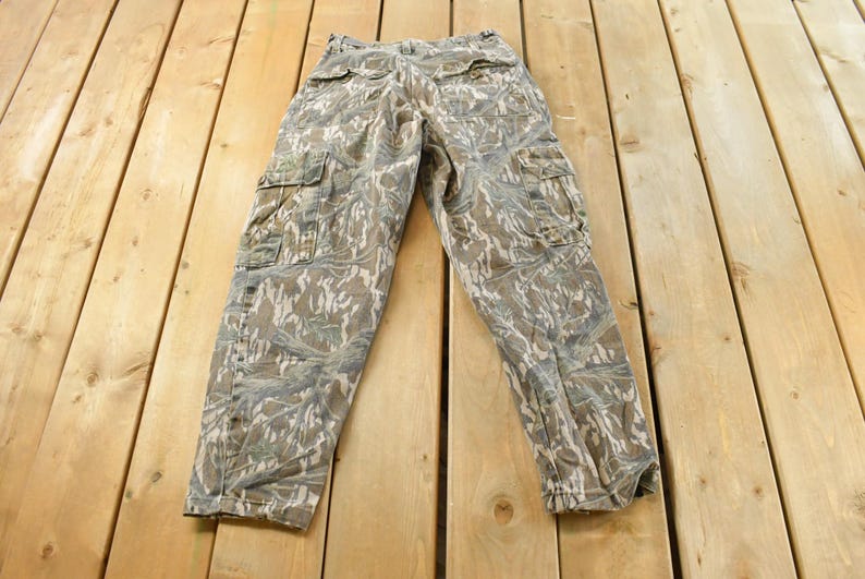 May include: Camouflage cargo pants with multiple pockets, in shades of brown, green, and gray. The pants are laid flat on a wooden surface, showcasing the detailed pattern and design.