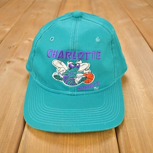 Op de afbeelding: Tealblauwe baseballcap met een geborduurd Charlotte Hornets-logo met een cartoon-hoornaar met een basketbal. De tekst "Charlotte Hornets TM" is geborduurd op de cap.