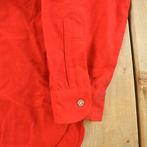 May include: Close-up of a bright red long-sleeved shirt. The cuff of the sleeve is visible, featuring a button closure. The fabric appears to be a soft, possibly flannel material. The shirt is on a light wood surface.