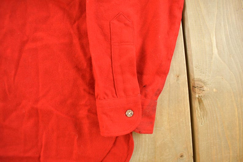 May include: Close-up of a bright red long-sleeved shirt. The cuff of the sleeve is visible, featuring a button closure. The fabric appears to be a soft, possibly flannel material. The shirt is on a light wood surface.