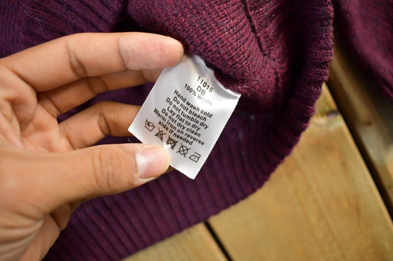 Puede incluir: Primer plano de un su&eacute;ter de lana burdeos con una etiqueta blanca. La etiqueta muestra las instrucciones de cuidado: lavar a mano con agua fr&iacute;a, no usar secadora, secar en plano, no limpiar en seco, planchar del rev&eacute;s con plancha tibia.