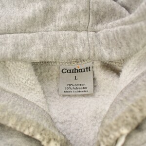 Puede incluir: Una sudadera con capucha gris Carhartt con una etiqueta blanca que dice "Carhartt" en letras negras, "L" en letras negras, "70% Algod&oacute;n 30% Poli&eacute;ster" en letras negras y "Hecho en M&eacute;xico" en letras negras.