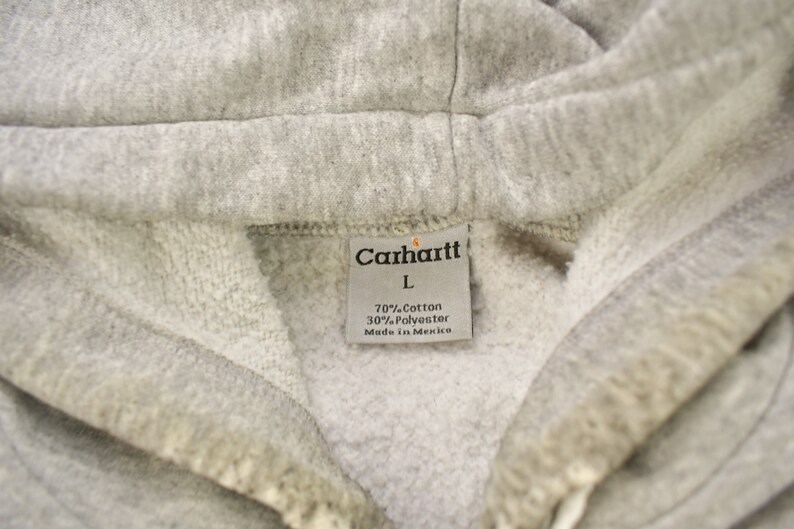 Puede incluir: Una sudadera con capucha gris Carhartt con una etiqueta blanca que dice "Carhartt" en letras negras, "L" en letras negras, "70% Algod&oacute;n 30% Poli&eacute;ster" en letras negras y "Hecho en M&eacute;xico" en letras negras.