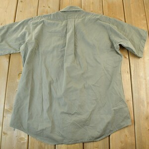 Peut inclure: Une chemise boutonn&eacute;e &agrave; manches courtes vert clair, vue de dos, est pos&eacute;e sur une surface en bois. La chemise a un col et une couture verticale au centre. Le tissu semble l&eacute;g&egrave;rement froiss&eacute;.