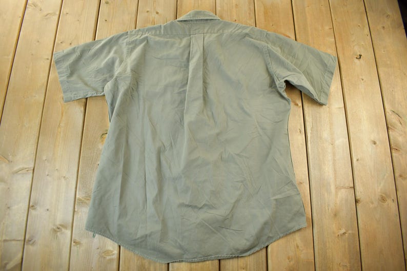Peut inclure: Une chemise boutonn&eacute;e &agrave; manches courtes vert clair, vue de dos, est pos&eacute;e sur une surface en bois. La chemise a un col et une couture verticale au centre. Le tissu semble l&eacute;g&egrave;rement froiss&eacute;.