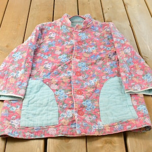 May include: A quilted jacket featuring a pink base with a floral pattern in shades of blue, yellow, and white. The jacket has a stand-up collar, long sleeves, a front button closure with red buttons, and two large, light blue patch pockets on the front.