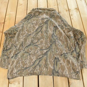 Op de afbeelding: Een camouflage shirt met lange mouwen en een hoge kraag. Het shirt heeft een patroon van bruine, bruine en grijze boomtakachtige ontwerpen. Het shirt ligt op een houten oppervlak.