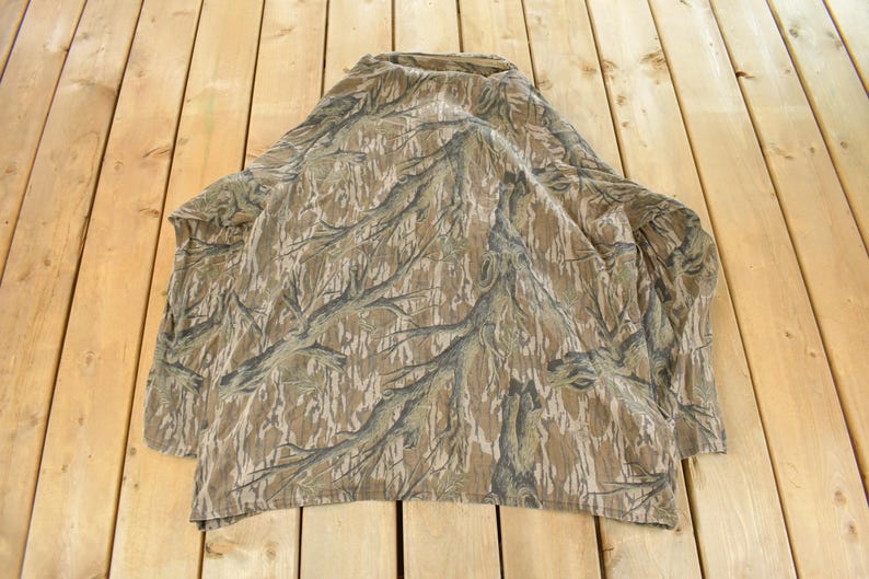Op de afbeelding: Een camouflage shirt met lange mouwen en een hoge kraag. Het shirt heeft een patroon van bruine, bruine en grijze boomtakachtige ontwerpen. Het shirt ligt op een houten oppervlak.