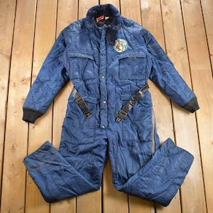 May include: A blue quilted one-piece jumpsuit with a zipper and snap closure. The jumpsuit has a black belt with buckles and a patch on the chest with a logo.
