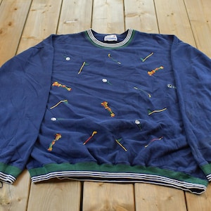 May include: A navy blue long-sleeve sweatshirt featuring an all-over embroidered pattern of golf clubs, golf balls, and flags. The garment has a crew neckline and ribbed cuffs and hem, both accented with horizontal green and white stripes.
