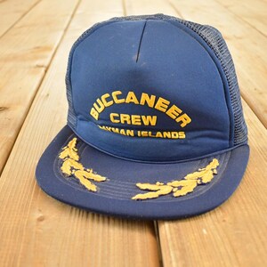 Puede incluir: Gorra de camionero azul marino con la parte trasera de malla. La parte delantera de la gorra presenta las palabras "BUCCANEER CREW CAYMAN ISLANDS" en letras doradas. Diseños dorados en forma de hojas están bordados en la visera.