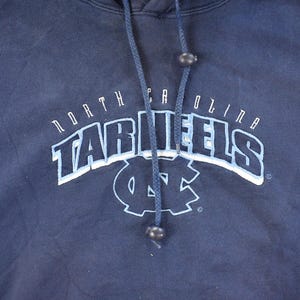 K&ouml;nnte beinhalten: Marineblauer Kapuzenpullover mit den Worten "North Carolina Tar Heels" in wei&szlig;er und hellblauer Bogenbeschriftung. Das Logo der University of North Carolina befindet sich unter dem Text. Der Pullover hat einen Kordelzug und eine Kapuze.