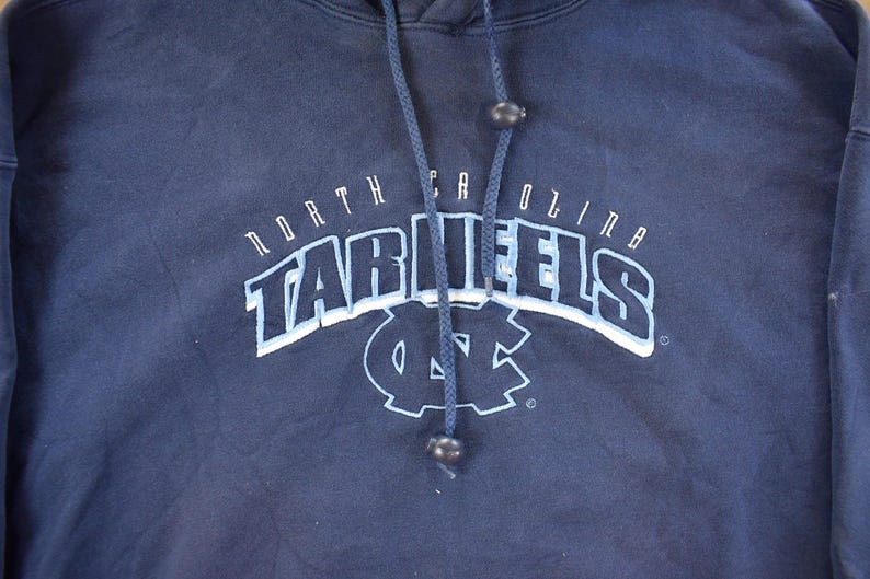 K&ouml;nnte beinhalten: Marineblauer Kapuzenpullover mit den Worten "North Carolina Tar Heels" in wei&szlig;er und hellblauer Bogenbeschriftung. Das Logo der University of North Carolina befindet sich unter dem Text. Der Pullover hat einen Kordelzug und eine Kapuze.