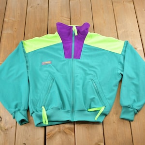 Puede incluir: Una chaqueta azul turquesa con cremallera con detalles en verde neón y morado. La chaqueta tiene cierre de cremallera y dos bolsillos. El logotipo de Columbia es visible en la parte delantera de la chaqueta.