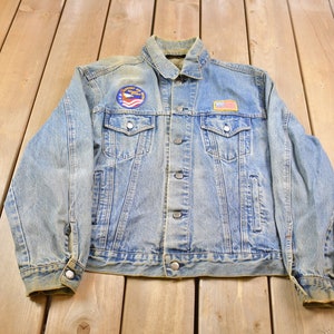 Pode incluir: Uma jaqueta de ganga azul claro com um visual desbotado. Ela tem um remendo no peito esquerdo com as palavras "United States" e uma pequena bandeira americana. Há um remendo menor no peito direito com um design vermelho, branco e azul.