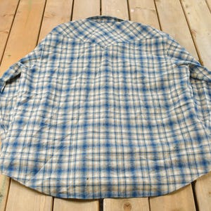 May include: A blue and white plaid button-down shirt. The shirt is made of a soft fabric and features a classic plaid pattern. The shirt is laid flat on a wooden surface.