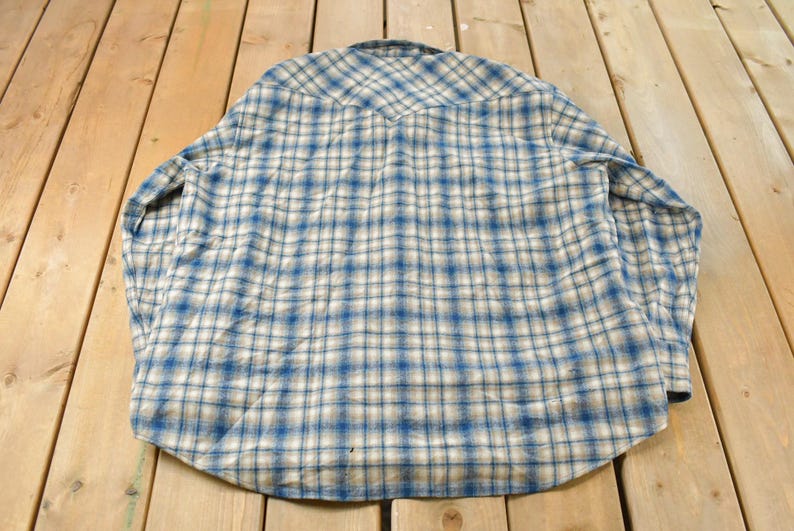 May include: A blue and white plaid button-down shirt. The shirt is made of a soft fabric and features a classic plaid pattern. The shirt is laid flat on a wooden surface.