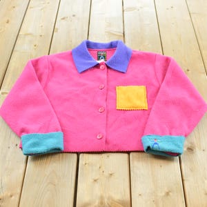 Könnte beinhalten: Eine leuchtend pinke Fleecejacke mit violettem Kragen, einer quadratischen gelben Brusttasche und türkisfarbenen Ärmelbündchen. Die Jacke hat eine durchgehende Knopfleiste und ein kleines grafisches Logo auf dem Innenetikett sowie am rechten Ärmel.