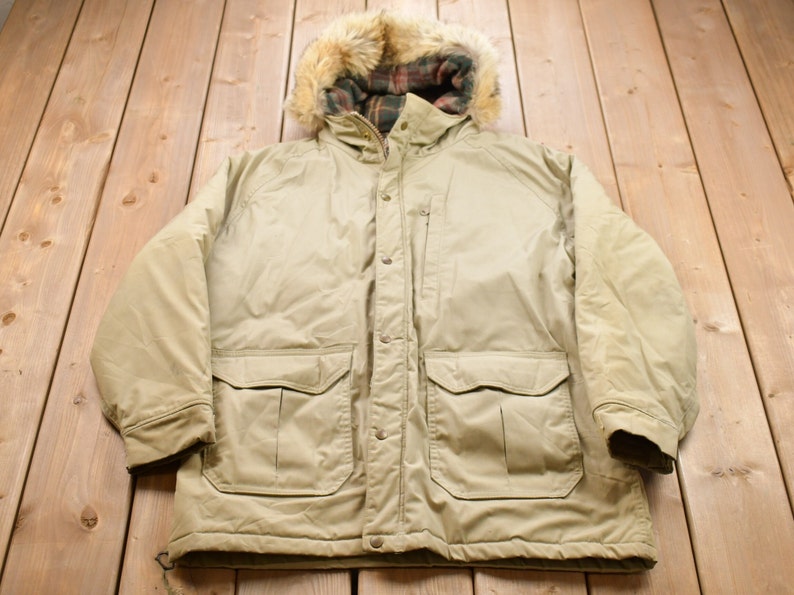 May include: A beige hooded winter parka with a plaid lining and two large flap pockets. The jacket has a zipper and button closure.