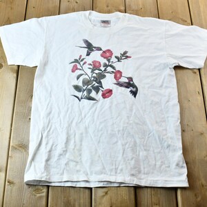 Peut inclure: T-shirt blanc avec un imprimé de colibris et de fleurs roses.