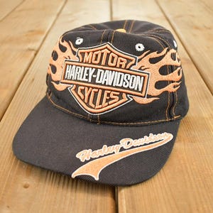 Może przedstawiać: Czarna czapka z daszkiem z logo Harley-Davidson Cycles w kolorze pomarańczowym i białym, otoczonym pomarańczowymi płomieniami. Daszek czapki ma napis Harley-Davidson w kolorze pomarańczowym. Akcesorium motocyklowe.