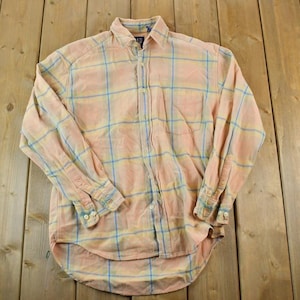 Könnte beinhalten: Ein langärmeliges Button-Down-Hemd mit Karomuster. Das Hemd hat eine pfirsichfarbene Basis mit einem hellblauen und gelben Gitterdesign. Es hat einen Kragen und geknöpfte Manschetten. Das Gap-Markenetikett ist sichtbar.