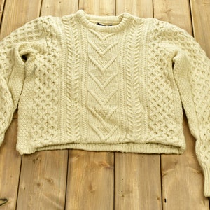 May include: A cream-colored cable knit sweater with a classic crew neck. The sweater is made of a soft, warm material and has a relaxed fit.