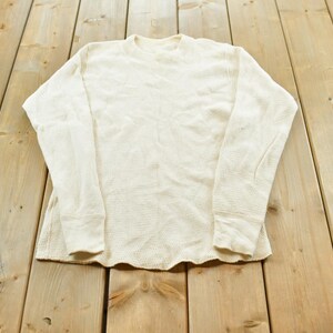 Peut inclure: Un haut thermique à manches longues de couleur crème blanc cassé. Le vêtement présente une encolure ronde et un motif en tissu gaufré texturé sur tout le corps et les manches, avec des poignets côtelés et un ourlet droit au bas.