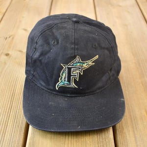 May include: Black baseball cap with an embroidered logo of a marlin fish in green, blue and gold, with the letter "F" on its side.