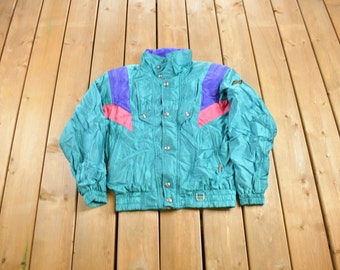 Könnte beinhalten: Eine tealblaue Skijacke mit pinkfarbenen und violetten Akzenten. Die Jacke hat einen Reißverschluss und eine Druckknopfleiste.