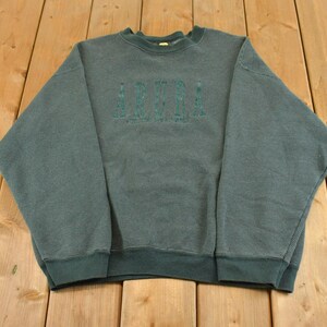 May include: A dark forest green crewneck sweatshirt featuring a textured, ribbed fabric throughout. The word ARUBA is embroidered in a darker green thread across the center of the chest in a bold, block-style font with a subtle shadow effect.