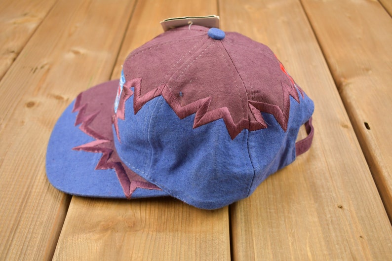 May include: A blue and burgundy baseball cap with a zig-zag pattern around the brim. The cap has a burgundy crown and a blue bill.