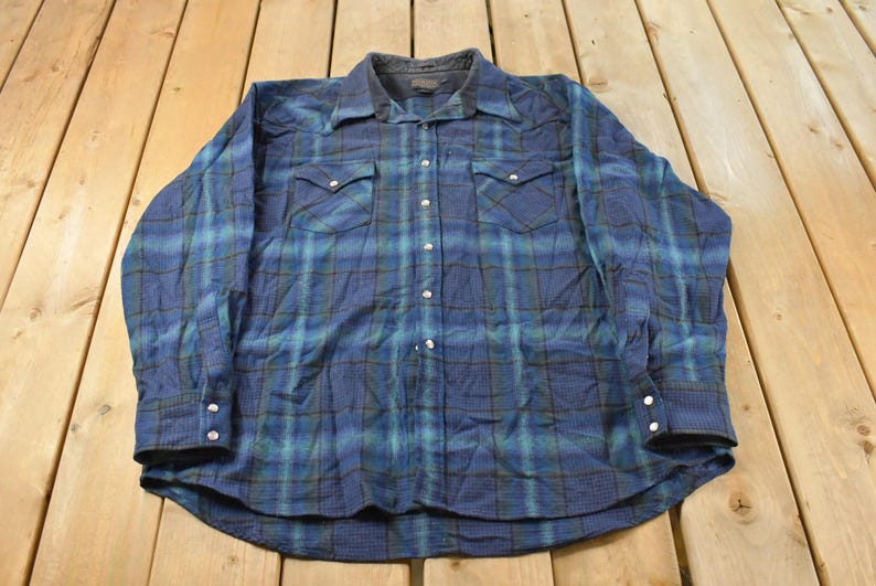 May include: Blue and green plaid button-down shirt with two chest pockets and snap closures. The long-sleeved shirt has a dark collar and cuffs. The shirt is made of a soft, comfortable material.