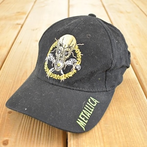Op de afbeelding: Zwarte baseballcap met een geborduurd schedelontwerp en een gele halo. Het woord "METALLICA" is in het groen op de zijkant gedrukt. De cap is gemaakt van een stevige stof en heeft een gebogen klep.