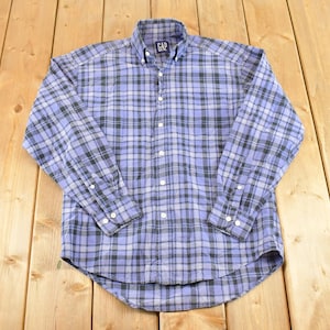 Könnte beinhalten: Ein blau-schwarz kariertes Hemd mit langen Ärmeln und Knopfleiste. Das Hemd hat einen Button-Down-Kragen und Manschetten. Der Markenname "GAP" ist auf dem Kragen sichtbar. Das Hemd liegt flach auf einer Holzoberfläche.