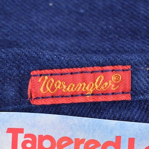 May include: A red label with the word "Wrangler" in gold lettering on a dark blue denim background.