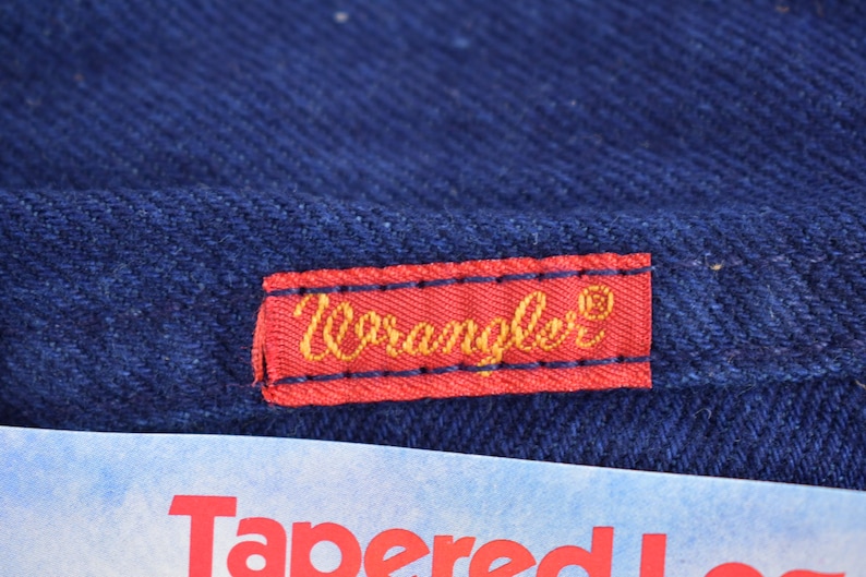May include: A red label with the word "Wrangler" in gold lettering on a dark blue denim background.