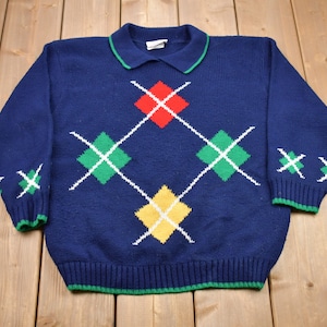 May include: Navy blue sweater with a collared neckline and argyle pattern. The argyle design features red, green, and yellow diamonds outlined in white. The collar and cuffs have green trim.