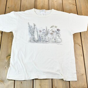 以下が含まれることがあります： ガラス瓶に花が描かれた白いTシャツ。絵はグレーの濃淡で描かれており、Tシャツは柔らかく快適な生地で作られています。