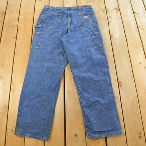 K&ouml;nnte beinhalten: Blaue Jeans-Arbeitshose mit mehreren Taschen, darunter eine Seitentasche und Ges&auml;&szlig;taschen. Die Hose liegt flach auf einer Holzoberfl&auml;che und zeigt ihr Design und ihre Konstruktion. Die Jeans sind ein klassischer Arbeitskleidungsstil.