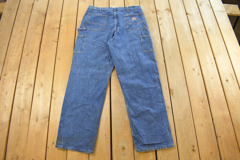 K&ouml;nnte beinhalten: Blaue Jeans-Arbeitshose mit mehreren Taschen, darunter eine Seitentasche und Ges&auml;&szlig;taschen. Die Hose liegt flach auf einer Holzoberfl&auml;che und zeigt ihr Design und ihre Konstruktion. Die Jeans sind ein klassischer Arbeitskleidungsstil.