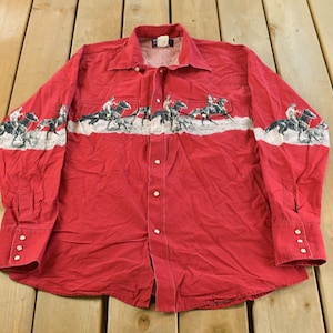 Puede incluir: Camisa roja con botones y una banda blanca con una ilustración en blanco y negro de vaqueros a caballo. La camisa de manga larga tiene cuello y puños abotonados.