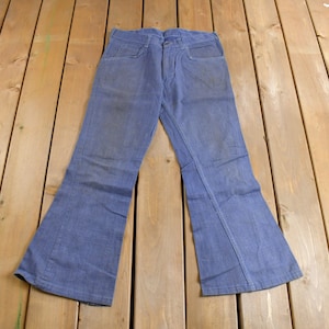 Pode incluir: Calças de ganga azul claro com design de perna larga. As calças de ganga têm um estilo clássico de cinco bolsos e um fecho de botão e zíper. As calças estão estendidas numa superfície de madeira.