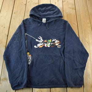 May include: Navy blue hooded sweatshirt featuring a cartoon graphic of Bugs Bunny, Daffy Duck, Marvin the Martian, and other Looney Tunes characters. The sweatshirt has a front pocket and a small Tweety Bird patch with a "B" logo.