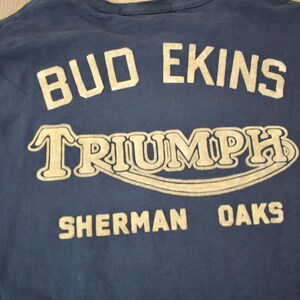 May include: A navy blue long-sleeved t-shirt with a gold print that reads "Bud Ekins Triumph Sherman Oaks".