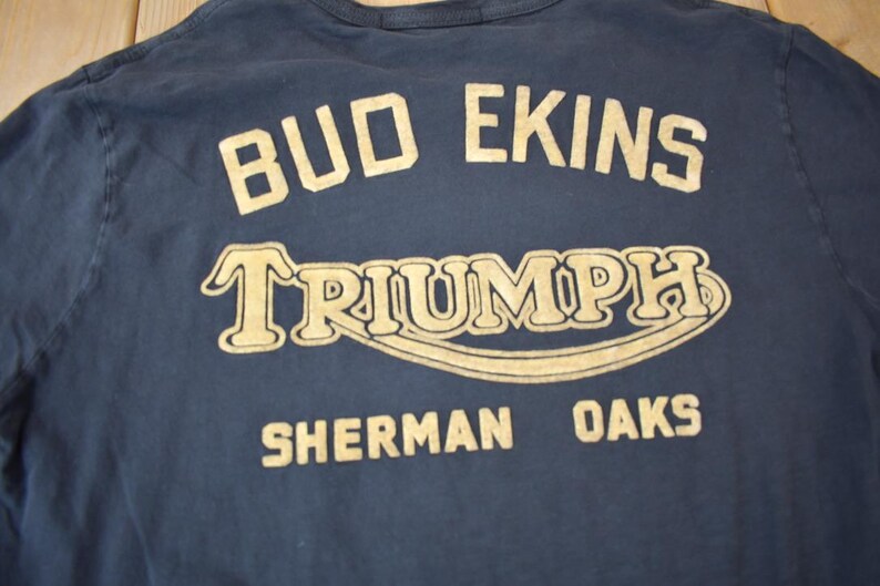 May include: A navy blue long-sleeved t-shirt with a gold print that reads "Bud Ekins Triumph Sherman Oaks".