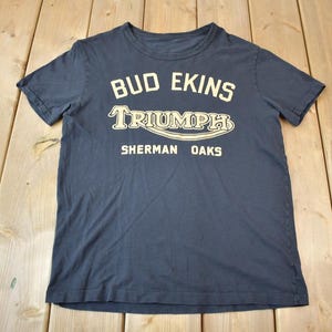 May include: A dark blue t-shirt with a white graphic print that reads "Bud Ekins Triumph Sherman Oaks".
