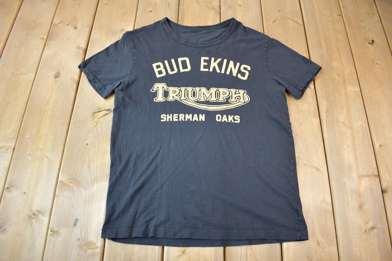 May include: A dark blue t-shirt with a white graphic print that reads "Bud Ekins Triumph Sherman Oaks".