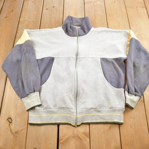 Könnte beinhalten: Ein grauer und blauer Reißverschluss-Sweatshirt mit gelbem Besatz. Der Sweatshirt hat einen Stehkragen und zwei Taschen.