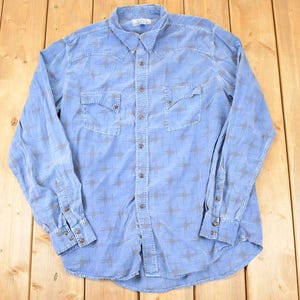 May include: Light blue button-down shirt with long sleeves and two chest pockets. The shirt features a subtle pattern of small, gray starburst designs. The collar and cuffs are a slightly darker shade of blue.